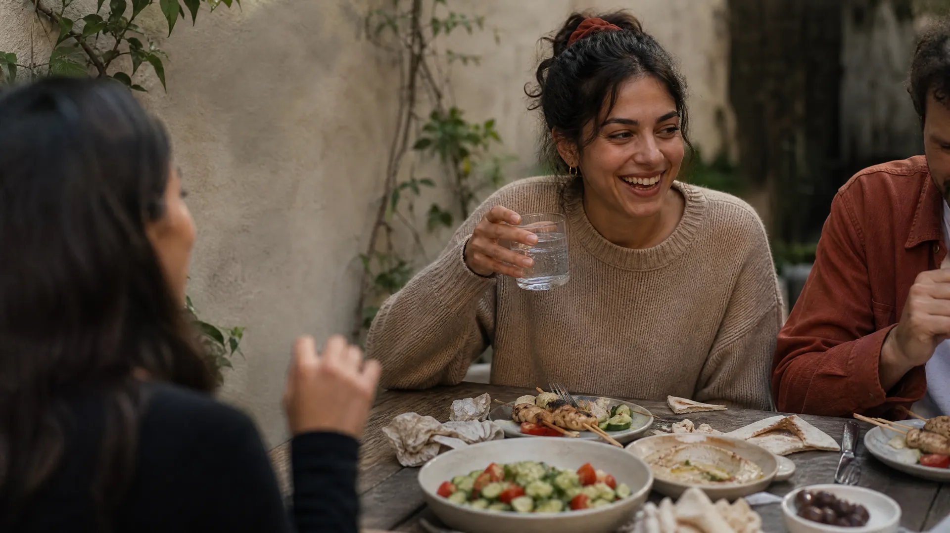 A woman laughs with friends over a shared table of chicken skewers, salad, hummus and flatbread in a courtyard garden.